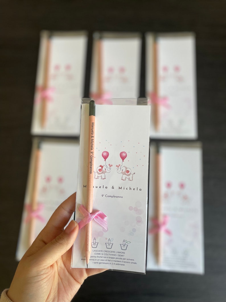 May include: Two pink and white elephant illustrations with balloons above them. The text "Manuela & Michela 9' Compleanno" is printed on the card. A wooden pencil with a pink ribbon is attached to the card.