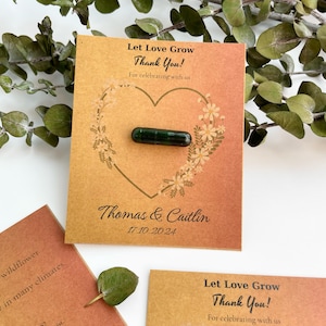Può includere: Un biglietto marrone con il testo "Let Love Grow Thank You!" e "For celebrating with us". Il biglietto ha la forma di un cuore con un pacchetto di semi verde al suo interno. Il testo "Thomas & Caitlin" e "17.10.2024" è stampato sotto il cuore.