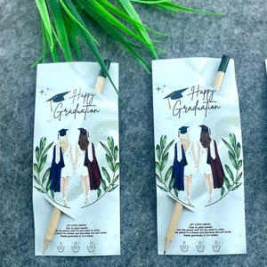 Może przedstawiać: Dwie karty z okazji ukończenia studi&oacute;w z oł&oacute;wkiem, kt&oacute;ry jest r&oacute;wnież paczką nasion. Karty przedstawiają rysunek dw&oacute;ch kobiet w togach i czapkach absolwent&oacute;w. Tekst na kartach brzmi "Happy Graduation" i "Let Love Grow".