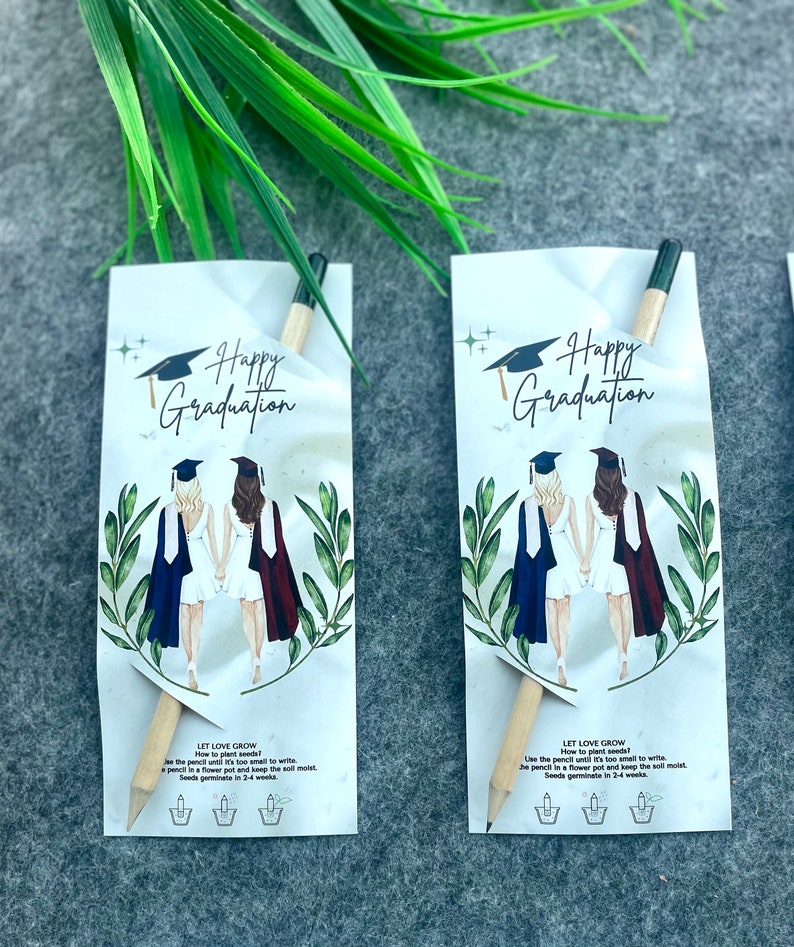 Może przedstawiać: Dwie karty z okazji ukończenia studi&oacute;w z oł&oacute;wkiem, kt&oacute;ry jest r&oacute;wnież paczką nasion. Karty przedstawiają rysunek dw&oacute;ch kobiet w togach i czapkach absolwent&oacute;w. Tekst na kartach brzmi "Happy Graduation" i "Let Love Grow".