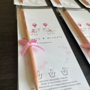 May include: A pink and white card with two pink elephants and the text "Muela & Michela 9 Compleanno". A wooden pencil with a pink ribbon is attached to the card.