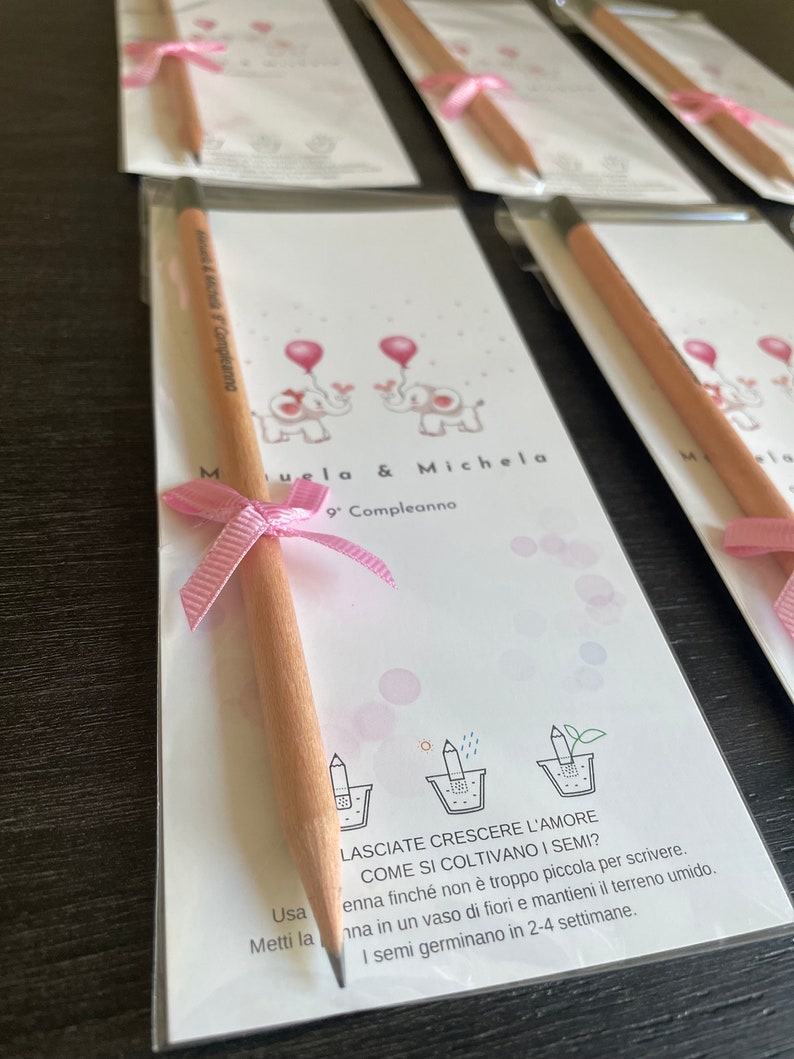 May include: A pink and white card with two pink elephants and the text "Muela & Michela 9 Compleanno". A wooden pencil with a pink ribbon is attached to the card.