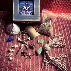 May include: A wooden box with a picture of a man in the ocean, surrounded by various sea-themed items, including seashells, an anchor, a piece of driftwood, and a small bag of dried herbs.
