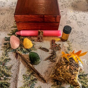 Puede incluir: Una colección de artículos espirituales que incluye una caja de madera, una vela rosa, cristales, pequeños viales, amuletos, una varita de madera y una bolsa de hierbas. Los artículos están dispuestos sobre una tela estampada.