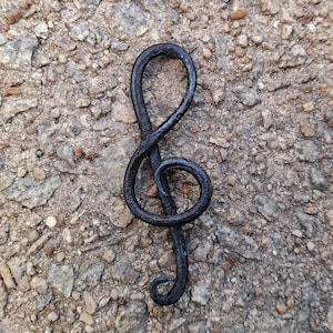 May include: A black metal treble clef music note charm. The charm is made of a single piece of metal that is bent into the shape of a treble clef. The charm is lying on a rough, gray surface.