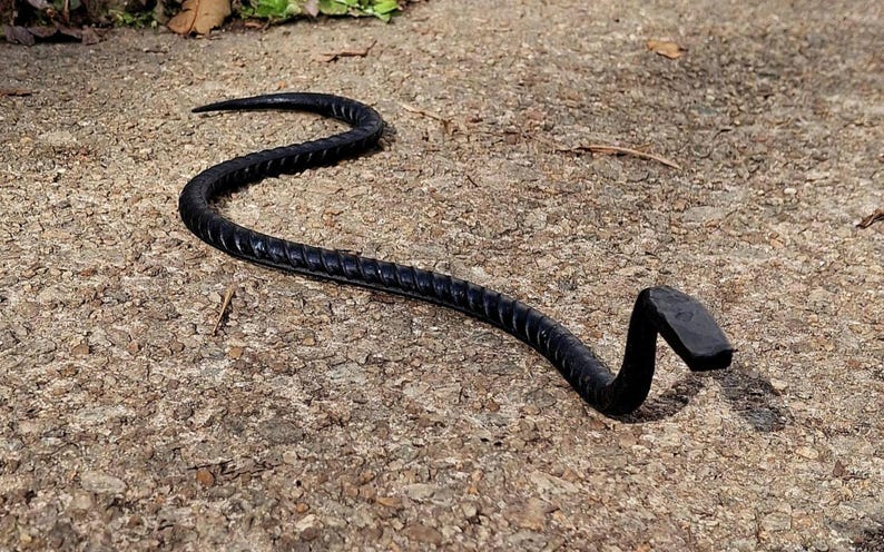 K&ouml;nnte beinhalten: Eine schwarze Schlangenskulptur aus Metall mit einem eingerollten Schwanz und einem flachen Kopf. Die Skulptur liegt auf einer Betonfl&auml;che.