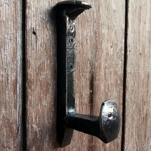 May include: A black metal hook shaped like a nail, mounted on a wooden surface. The hook is designed to hang items on a wall.