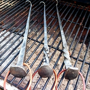 May include: Three metal grilling skewers with leather loops. The skewers are made of twisted metal and have a hook at the top. The skewers are on a black grill grate.