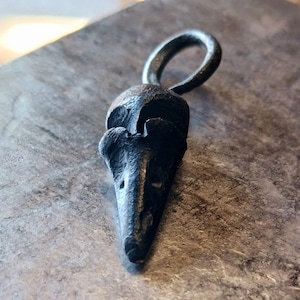 May include: A handcrafted, dark metal pendant in the shape of a bird's head, featuring a textured surface and a circular loop at the top. The pendant is likely made of iron or a similar metal, and it has a rustic, artisanal appearance.