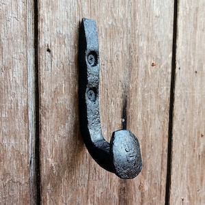 May include: Black metal hook with a rounded head, mounted on a wooden surface. The hook is made from a repurposed nail.