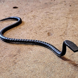 K&ouml;nnte beinhalten: Eine schwarze Schlangenskulptur aus Metall mit einem eingerollten Schwanz und einem flachen Kopf. Die Skulptur ist aus verdrehtem Metall gefertigt und hat eine raue, strukturierte Oberfl&auml;che.