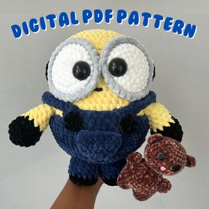 Op de afbeelding: Een gehaakt pluche speelgoed van een minion die een bruine teddybeer vasthoudt. De minion is geel met een blauwe overall en grote witte ogen. Het speelgoed is gemaakt van garen en heeft een vriendelijke uitdrukking.
