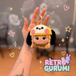 May include: A crochet keychain featuring a character with blonde hair and black ears, wearing a yellow hat with a white top and pink cheeks. The character has black bead eyes and is attached to a gold-toned metal lobster clasp and ring.