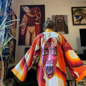 May include: A long-sleeved kimono-style jacket featuring a vibrant, hand-painted design on the back. The artwork depicts a large, stylised red devil face with horns and an open mouth, surrounded by abstract orange, yellow, and red wave patterns.