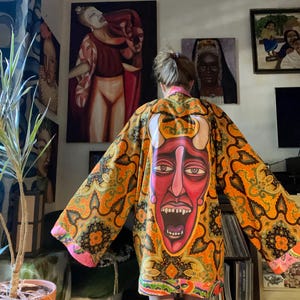 May include: A vibrant orange and yellow patterned kimono featuring a large red face design on the back. The kimono has long sleeves and pink edging. The person is standing in front of several paintings.