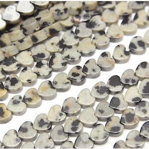 May include: Close-up of heart-shaped beads. The beads are a light beige color with black and gray spots, resembling a dalmatian's coat. The beads are strung together, suggesting they are for jewelry making or crafting.
