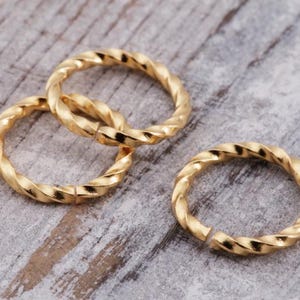 Op de afbeelding: Close-up van drie goudkleurige, gedraaide metalen ringen op een lichtgrijze houten ondergrond. Eén ring is los, terwijl twee in elkaar grijpen. De ringen hebben een getextureerd, touwachtig uiterlijk en reflecteren licht.