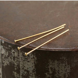 Peut inclure: Trois épingles à tête dorées sont disposées en diagonale sur une surface texturée brun foncé. Chaque épingle a une petite tête plate et une longue tige fine. Ces épingles sont probablement utilisées pour la fabrication de bijoux ou l'artisanat.