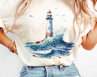 May include: A white t-shirt featuring a watercolour illustration of a lighthouse and crashing waves. The lighthouse is white with a red roof and is surrounded by blue and white waves.