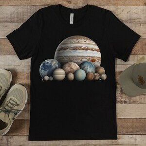 May include: A black t-shirt with a graphic of a group of planets, including Jupiter, Saturn, and Earth. The planets are arranged in a cluster, with Jupiter being the largest and most prominent.