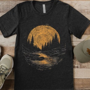 Puede incluir: Una camiseta gris oscuro con un estampado gráfico de una escena de bosque con un gran sol amarillo en el fondo. El sol tiene un patrón circular y el bosque está formado por árboles y un río.
