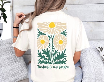 Puede incluir: Una camiseta blanca con un gráfico de tres margaritas estilizadas en tonos beige y verde. El texto "Tending to my garden" está impreso debajo del gráfico.