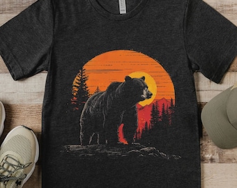 Può includere: Una maglietta grigio scuro con una stampa grafica di un orso nero in piedi di fronte a un tramonto con montagne e alberi sullo sfondo.