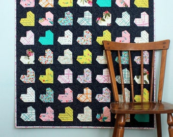 May include: A quilt with a black background and a patchwork design of colorful hearts. The quilt is hanging on a light blue wall with a wooden chair in front of it.