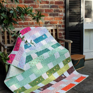May include: A colourful patchwork quilt featuring a range of floral and geometric patterns in shades of green, pink, orange, and blue. The quilt is folded and draped over a wooden bench.