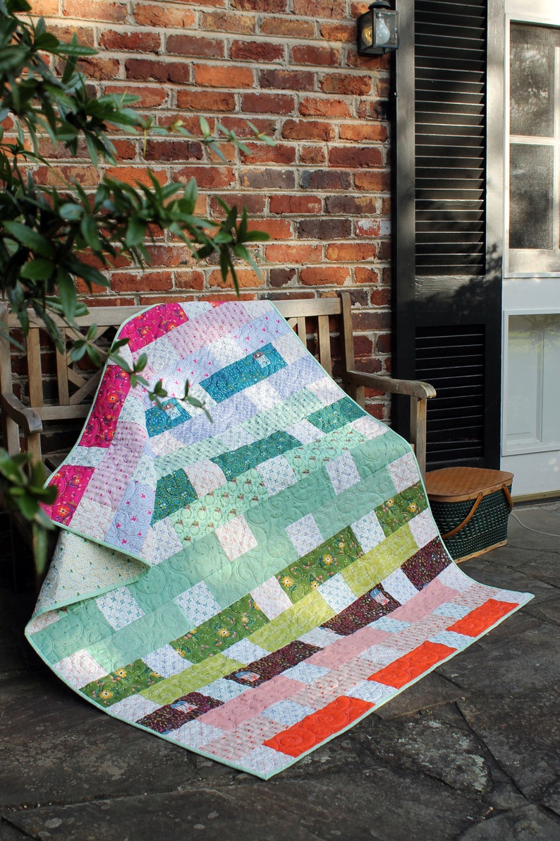 May include: A colourful patchwork quilt featuring a range of floral and geometric patterns in shades of green, pink, orange, and blue. The quilt is folded and draped over a wooden bench.