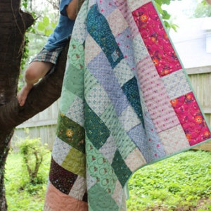 May include: A patchwork quilt with a variety of colours and patterns, including green, blue, pink, and white. The quilt is hanging from a tree branch.