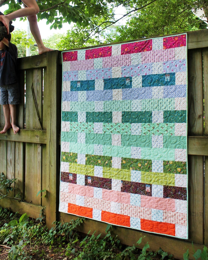 May include: A colourful patchwork quilt featuring a range of floral and geometric patterns in shades of pink, green, blue, and orange. The quilt is hanging on a wooden fence.