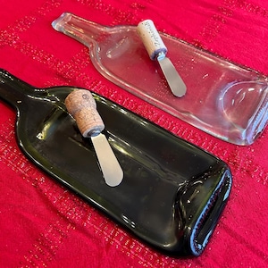 Puede incluir: Dos bandejas para servir con forma de botella, una transparente y otra verde oscuro, sobre un fondo rojo. Cada bandeja contiene un pequeño cuchillo para queso con mango de corcho. El mango de corcho de un cuchillo tiene el nombre "Zephaniah".