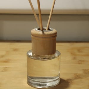 May include: A clear glass reed diffuser bottle filled with a transparent liquid, featuring a cylindrical light-colored wooden cap and three thin wooden reeds extending upward from the center opening of the bottle neck.