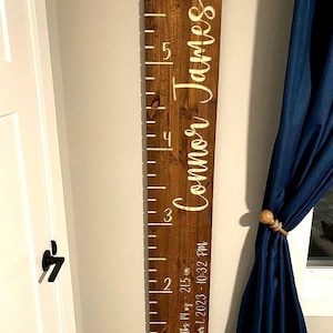 May include: A vertical wooden growth chart board with a dark brown stained finish. The board features white measurement markings and the name Connor James in script. Text includes 6 lbs 14 oz, 21.5 in, and November 1, 2023 - 10:32 PM.
