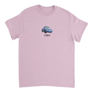 Op de afbeelding: Een lichtroze T-shirt met een cartoonafbeelding van een blauwe auto met een gezicht en het woord "Clairo" eronder gedrukt.