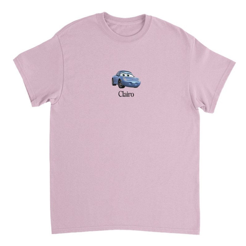 Op de afbeelding: Een lichtroze T-shirt met een cartoonafbeelding van een blauwe auto met een gezicht en het woord "Clairo" eronder gedrukt.