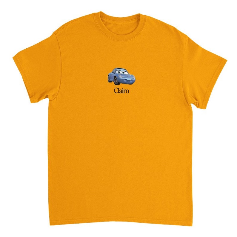Op de afbeelding: Een geel T-shirt met korte mouwen met een blauwe cartoonauto en het woord "Clairo" erop gedrukt.