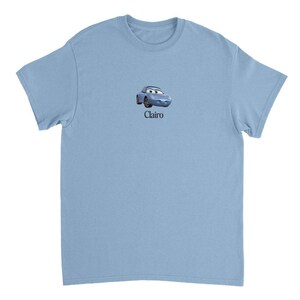 Op de afbeelding: Lichtblauw T-shirt met een cartoon afbeelding van een blauwe auto met een glimlachend gezicht en het woord "Clairo" gedrukt onder de afbeelding.