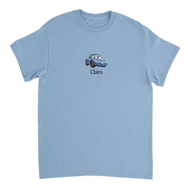 Op de afbeelding: Lichtblauw T-shirt met een cartoon afbeelding van een blauwe auto met een glimlachend gezicht en het woord "Clairo" gedrukt onder de afbeelding.