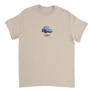 Op de afbeelding: Een licht beige t-shirt met een cartoon afbeelding van een blauwe auto met een glimlachend gezicht en het woord "Clairo" gedrukt onder de afbeelding.