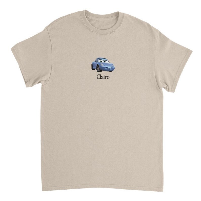 Op de afbeelding: Een licht beige t-shirt met een cartoon afbeelding van een blauwe auto met een glimlachend gezicht en het woord "Clairo" gedrukt onder de afbeelding.