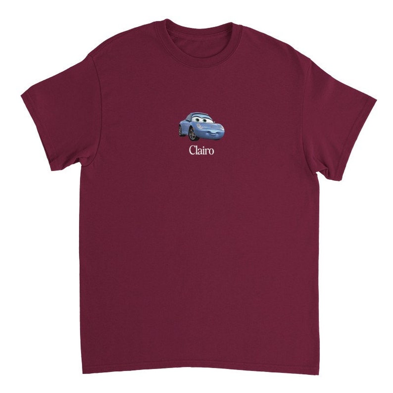 Op de afbeelding: Een maroon t-shirt met een cartoon afbeelding van een blauwe auto met een glimlach. De tekst "Clairo" is onder de auto gedrukt.