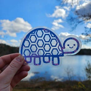 May include: A clear vinyl sticker of a blue and white turtle with a honeycomb pattern on its shell. The turtle has a smiling face and is holding its head up.