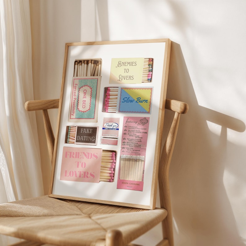 Puede incluir: Una obra de arte enmarcada con cajas de cerillas y tarjetas con textos como "Enemies to Lovers" y "Fake Dating". La obra de arte se exhibe en una silla de madera. La paleta de colores incluye rosa, azul y beige, creando una est&eacute;tica vintage.