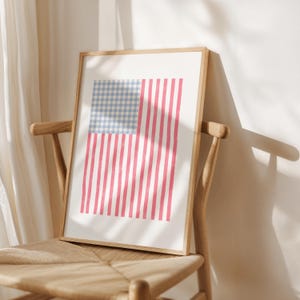 Puede incluir: Una impresión enmarcada de la bandera estadounidense. El diseño de la bandera presenta un patrón de cuadros azules y blancos en la esquina superior izquierda, con rayas verticales rojas y blancas. El marco es de madera clara.