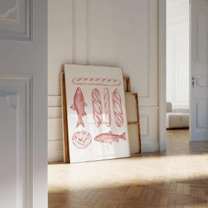 May include: A framed print featuring red illustrations of bread and fish. The artwork includes a baguette, rolls, and two fish, all rendered in a detailed, vintage style. The print is leaning against a white wall in a room with a wooden floor.
