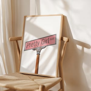 Puede incluir: Impresión enmarcada que muestra un letrero de calle rosa con las palabras "Pretty Girl Ave" en blanco. El letrero está fijado a un poste marrón y plateado. La impresión se muestra en una silla de madera.