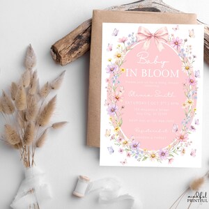 May include: A baby shower invitation with a pink oval frame, floral and butterfly accents, and the text "Baby in Bloom." The invitation is on a white background with a brown envelope.