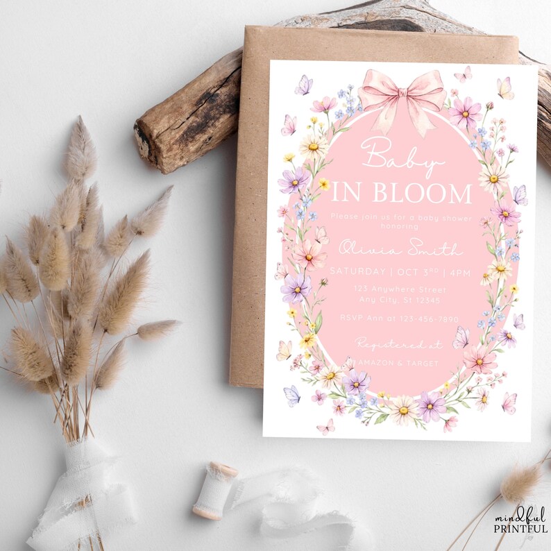 May include: A baby shower invitation with a pink oval frame, floral and butterfly accents, and the text "Baby in Bloom." The invitation is on a white background with a brown envelope.